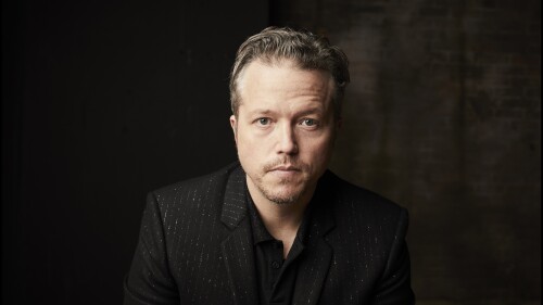 Musician Jason Isbell looks at the camera, wearing a black striped jacket in front of a dark background