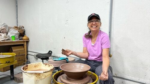 A widely grinning person sits before a completed bowl on a pottery wheel, hands wet with clay.