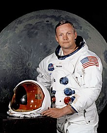A light-skinned man in his late 30s, with blue eyes and brown hair parted to the right. He wears a white space suit, and holds the helmet. The spacesuit has five hose connectors on the front. There is a large U.S. flag on the left shoulder. The helmet's transparent faceplate is tinted gold.