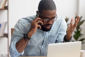 Angry mad african-american businessman talking on cellphone looking at laptop in office. Shocked manager received bad news discussing failing profit statistics with coworker, solving problem distantly