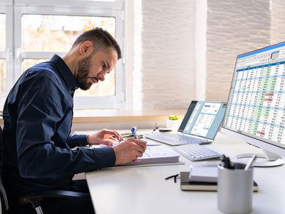 A man with a close-cropped beard seated at a desk writes on a notepad. Meanwhile a laptop screen and a desktop monitor display an invoice and a spreadsheet, respectively.