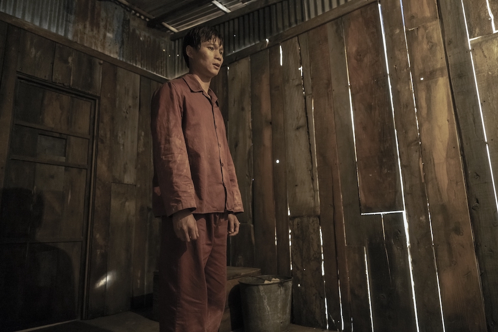 Hoa Xuande in The Sympathizer, shown here standing in a cell with wooden walls, wearing a plain brown uniform