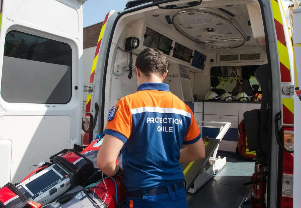 EMT in an orange and blue uniform stands outside the open doors of his ambulance