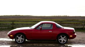 Red vintage Mazda Miata with an aftermarket hardtop coupe roof, parked in front of a green field.