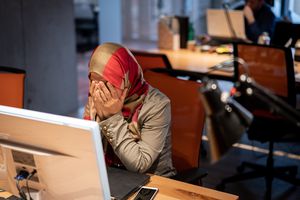 Woman wearing hijab, sitting at a computer desk with her hands covering her face