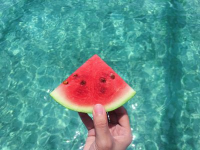 A White hand holding a slice of water melon over blue pool water.