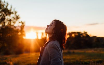 Side portrait of young woman with eyes closed inhaling fresh air, against sunset in the sky