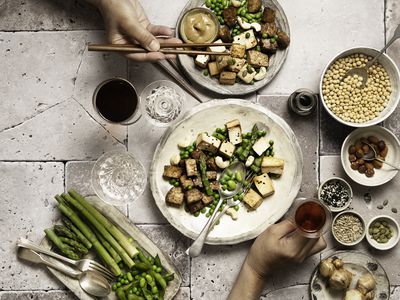 vegan and vegetarian foods in plates and bowls with human hands holding glasses, and different bowls containing plant based protein sources, miso sauce, seeds, asparagus, edamame, peas