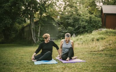 Smiling woman assisting friend while exercising on mat in public park - stock photo