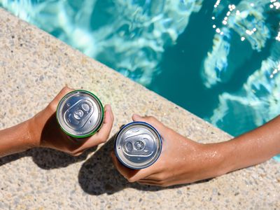 Two hands hold cans of cold drink in the pool on a sunny day.