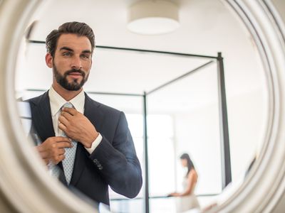 A man in a suit and tie looks admiringly at himself in a hotel room mirror. 