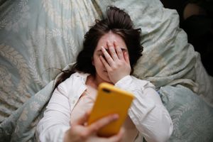 Young woman lying on a bed, covering her face with one hand, while holding a yellow cell phone in the other hand above her head. 