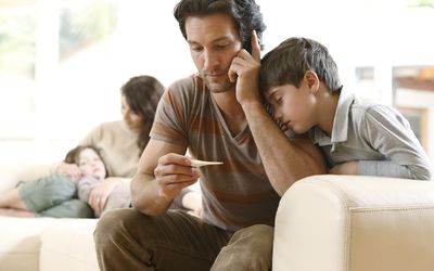 Man on the phone looking at a thermometer with his sick family in the room