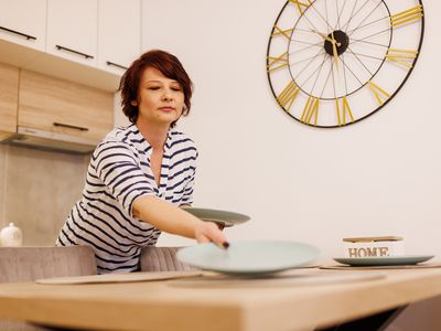 Low angle view of middle aged woman putting on plates while setting the table in front of large clock