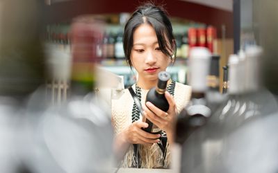 Young woman standing in a store, looking at the label of a wine bottle in her hands.