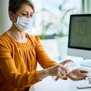 Woman in mask putting hand sanitizer on her hands at her desk