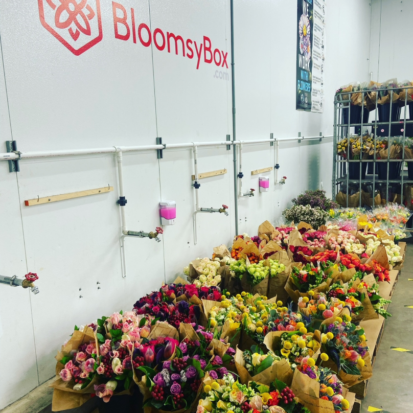 A large collection of colorful, flower bouquets beside a wall featuring the Bloomsybox logo