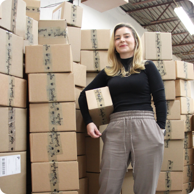 Jenni-Lyn Williams, CEO at SnarkyTea, holding a box for shipping and standing near stacks of cardboard boxes