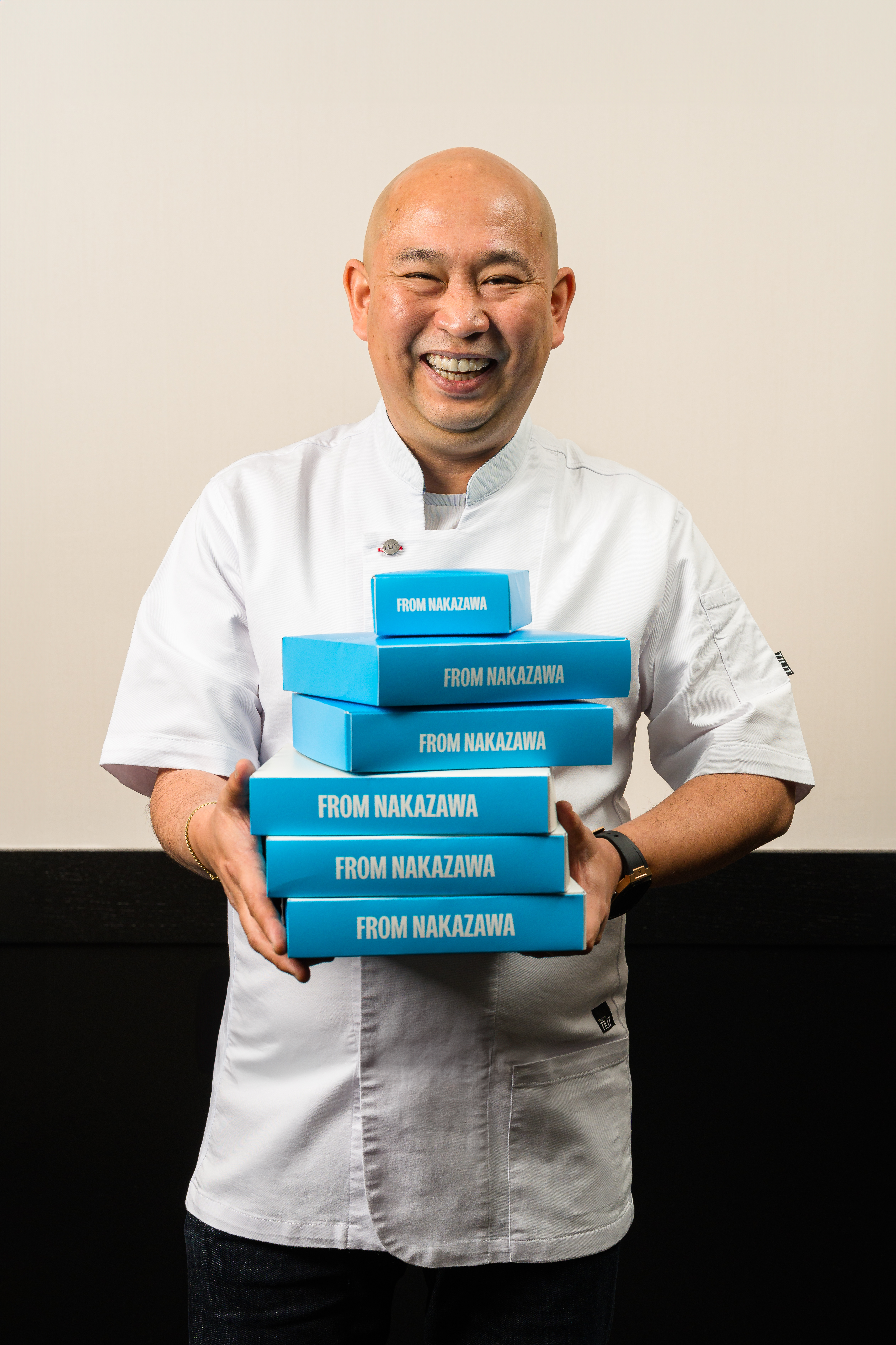 A smiling man in a white shirt carrying a stack of blue boxes that read Nakazawa.