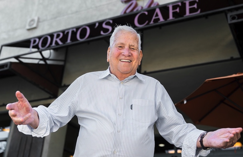 An elderly Cuban man stands with arms open in front of his bakery in Glendale, California called Porto’s Bakery.