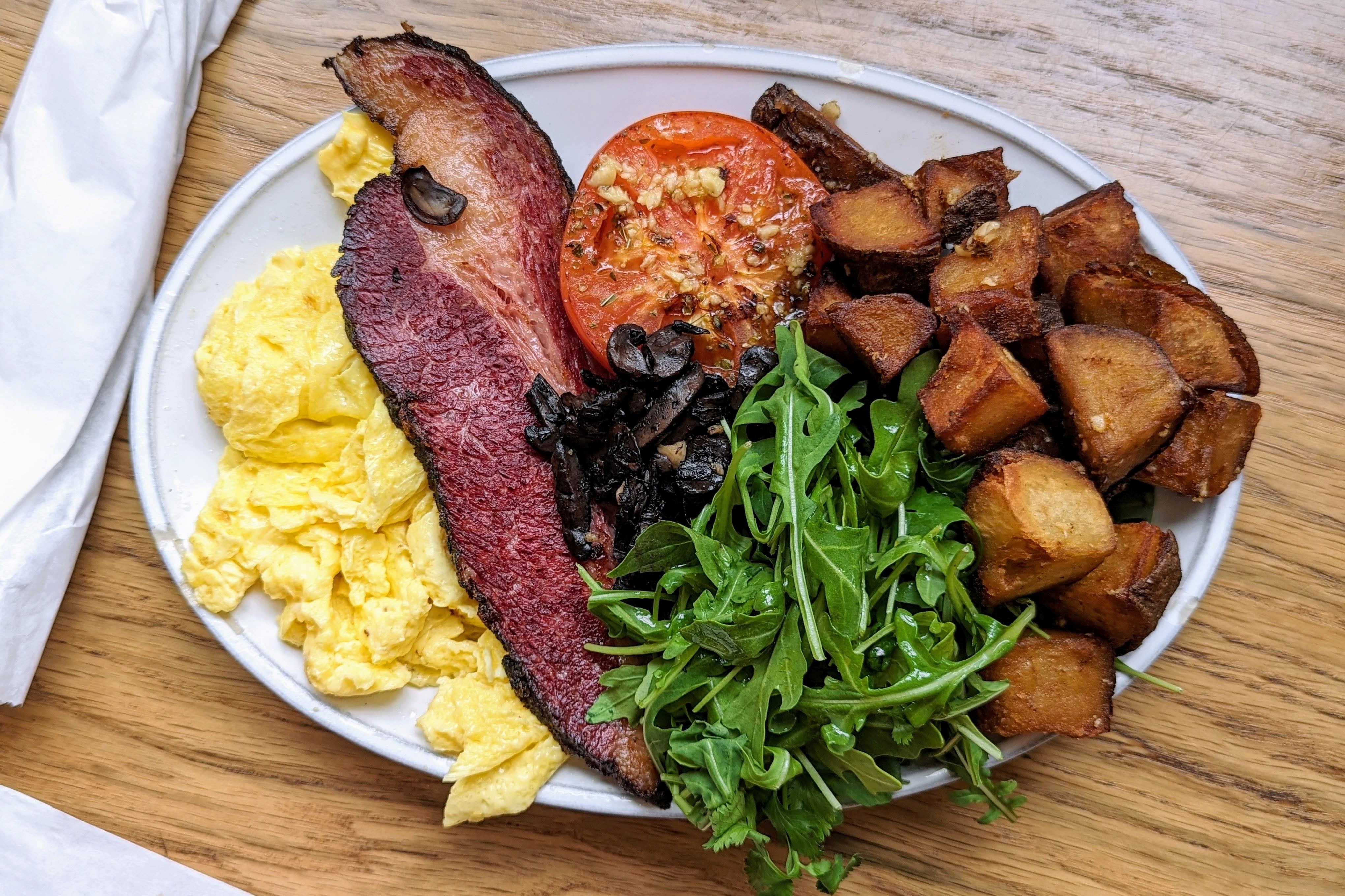Breakfast plate with scrambled eggs, bacon, arugula, mushrooms, tomato, and potatoes