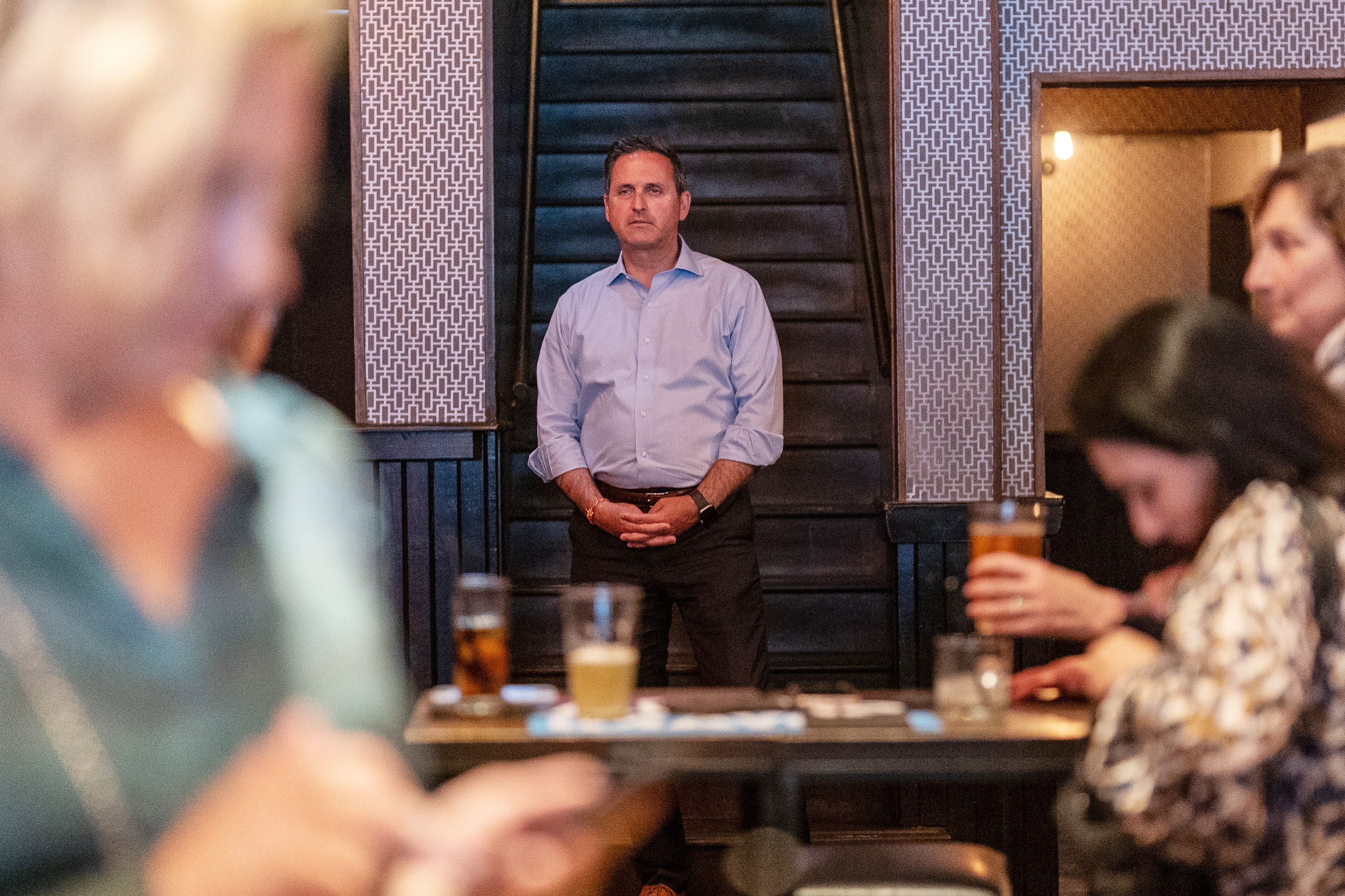 A man stands alone with a serious expression in a bustling bar scene, observed by unfocused patrons engaged in conversations and drinking.