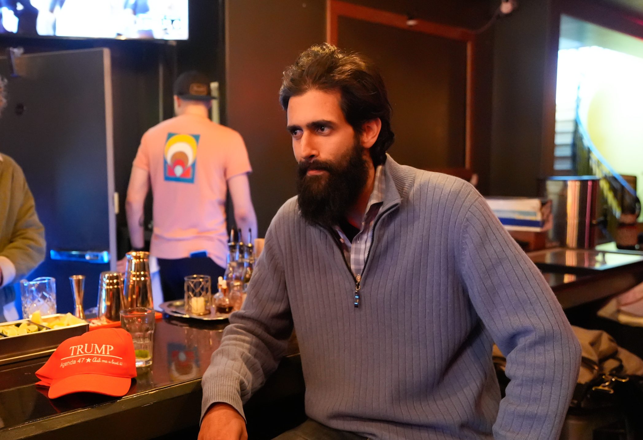 A man with a beard in a gray sweater is sitting at a bar counter, next to a red "Trump" hat. A bartender is preparing drinks and a TV is on in the background.