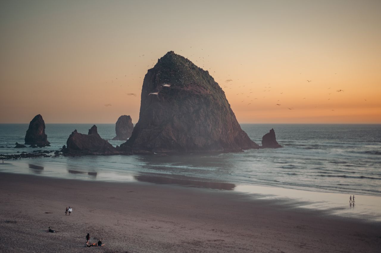 Sunset at Cannon Beach, Oregon, showing the towering, rugged Haystack Rock standing in the water, surrounded by smaller sea stacks, flying birds, and people on the sandy beach.