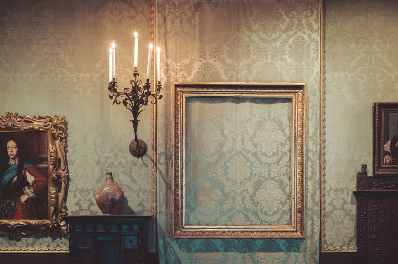 An empty picture frame in a room with ornate wallpaper, candles, old paintings, and antique furniture.