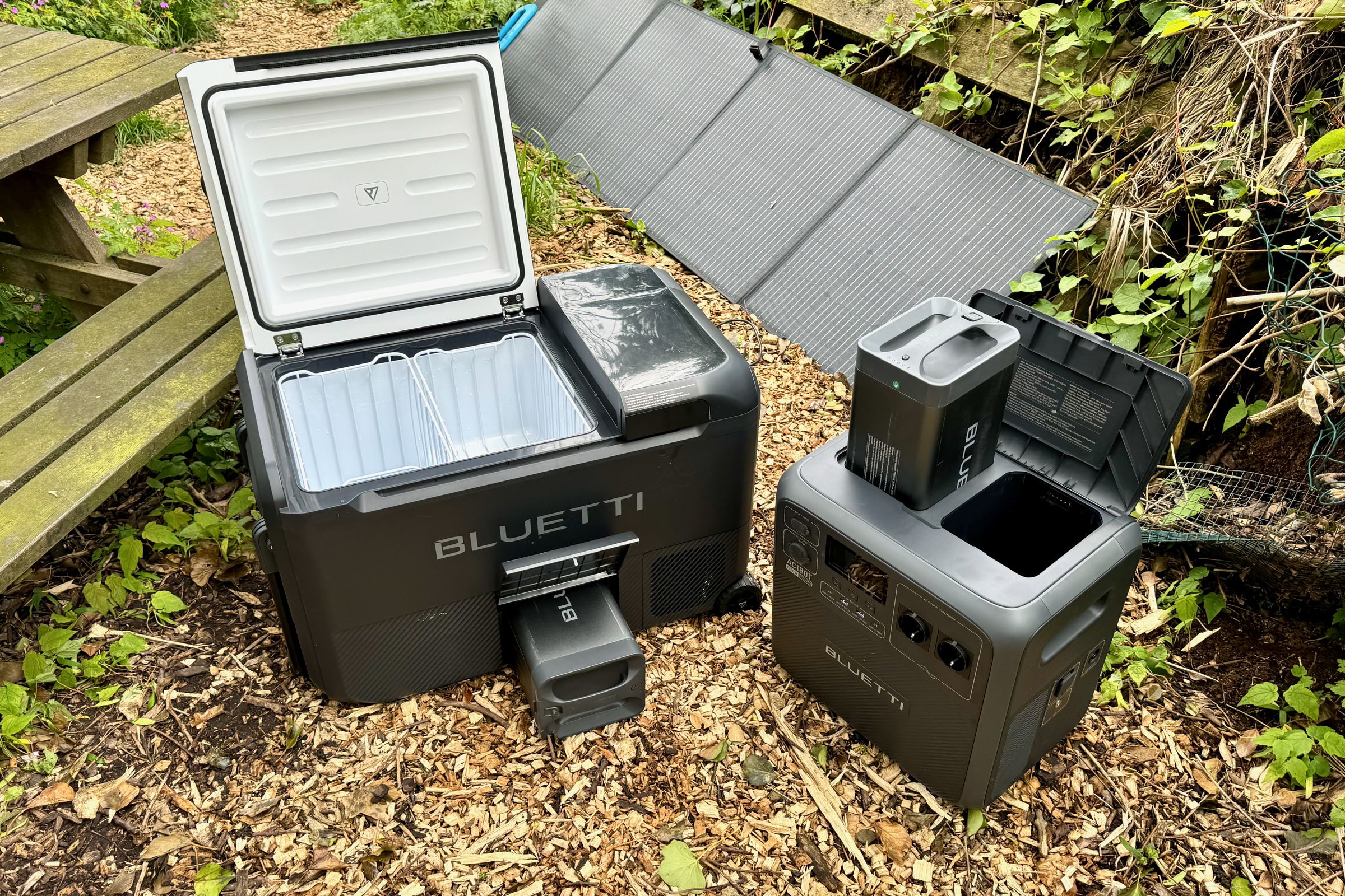 The Bluetti MultiCooler and AC180T solar generator sitting in a park next to a picnic table with a solar panel in the background.