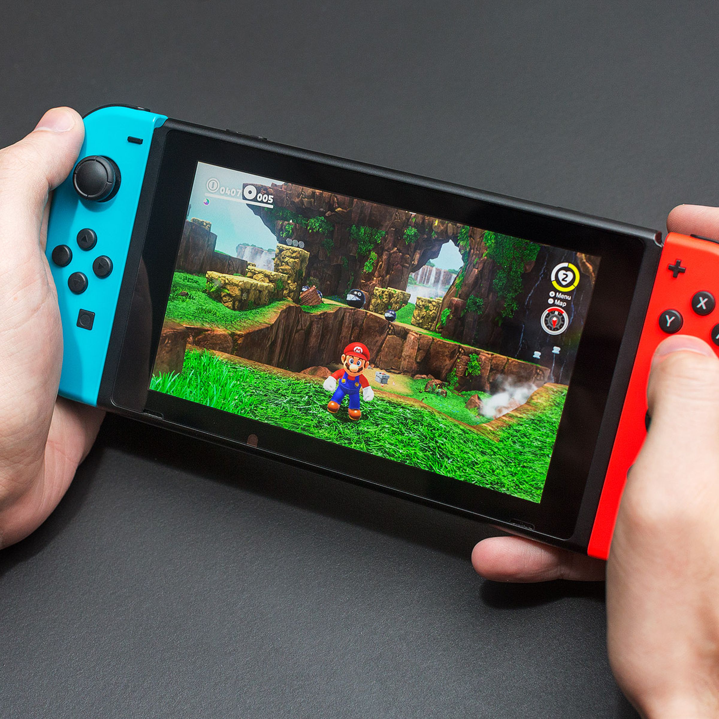 A pair of hands holding a Nintendo Switch console with neon red and blue Joy-Con controllers, playing Super Mario Odyssey in handheld mode.
