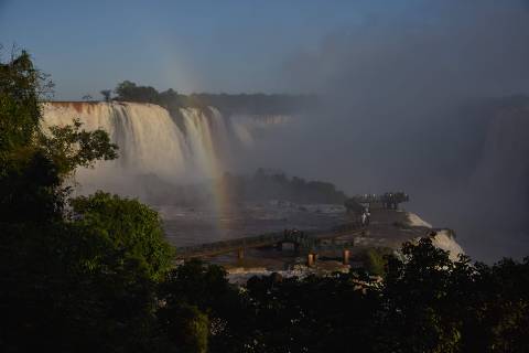 TOPSHOT - Visitors walks along a walkway as a rainbow appears in front of a partial view of the Devil's Throat Falls, part of the Iguazu Falls waterfall system on the Iguazu River at Iguacu National Park, Parana State, near Foz do Iguacu in southern Brazil on the border with Argentina, during sunset on May 21, 2024. The Iguazu Falls are shared by Brazil and Argentina. (Photo by Mariana SUAREZ / AFP)