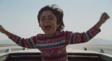 A young Iranian boy in a multicolored sweater with arms outstretched as he juts out of the roof of an automobile