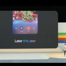 A woman stands in front of a large screen that displays the new Gemini app and reads "later this year."