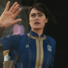 Lucy from "Fallout" steps out of the Vault and raises her hand to protect herself from the sun.