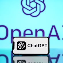 The ChatGPT logo is seen displayed on a mobile phone screen with OpenAI logo in the background.