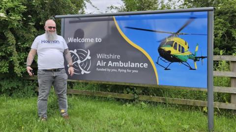 Picture shows Drew standing next to the Wiltshire Air Ambulance sign outside the charity's base. 