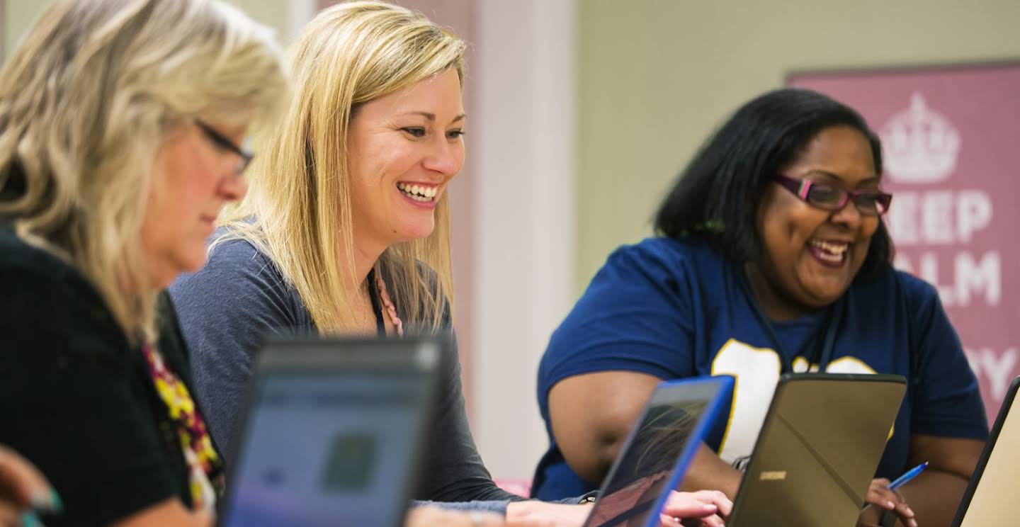 Tre kvinnelige lærere smiler foran datamaskinene sine.