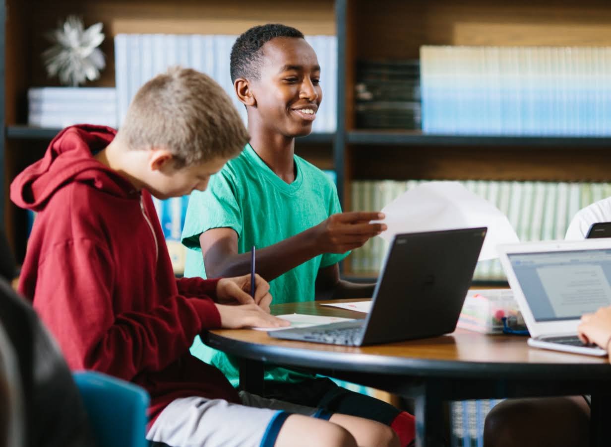Un alumno sostiene una hoja de papel sentado junto a otro alumno que trabaja frente a un Chromebook.