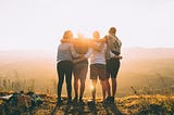 Four friends with arms around each other, facing a bright, sun-lit horizon