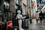 An empathetic looking, pristine white robot with a tablet interface offers its services  for sale in the alley of a mall.