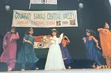 A 5-year-old Indian girl wears a white dress, tiara, and sash as she stands onstage. Surrounding her are other Indian girls, dancing in salwaar kameez outfits of differing colors.