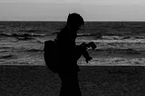 A black-and-white photo from Hanying Xie’s photobook, Behind the Camera. Photo of a man with a camera, silhouetted against a beach. Waves cascade onto the sand behind him. He stares down at his camera’s screen, contemplating his shots.