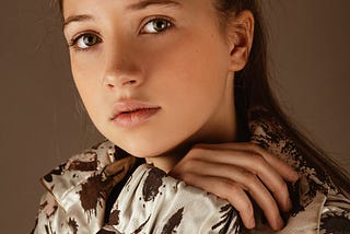 Young teenage girl with brown hair and light eyes stares at the Photographer Head shot. Girl is not smelling, and wears an artistic brown and white jacket.