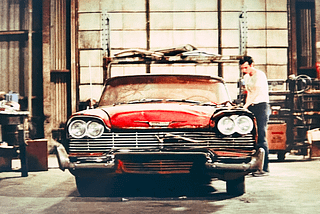 A red 1958 Plymouth Fury car which has a cracked windshield and various dents and broken body panels. There is a man getting into the car. It is a scene from the movie, Christine.