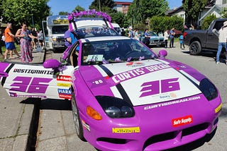 A purple and white sports car parked on the side of a road with people in the background.