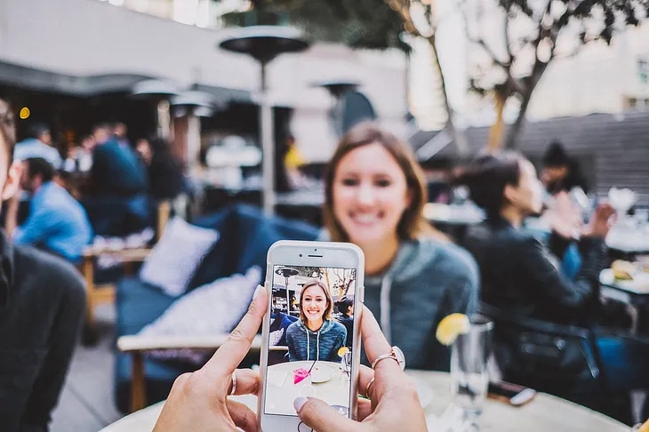 Person taking a photo with a smartphone of a young, smiling white woman who appears to be sitting at a crowded outdoor restaurant with heat lamps at each table. Her image is slightly blurry, except for the one on the phone.