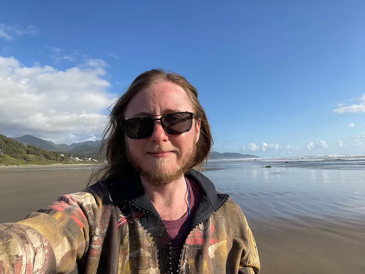 The author is standing between a beach and some mountains. He’s wearing a colorful jacket and sunglasses.