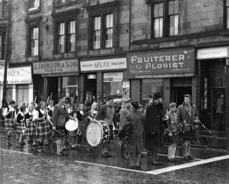 1960s photograph in black and white, showing a marching band in Scots dress in front of 4 shops on Maryhill Road, Glasgow, including Marion’s fish and chip shop and R. Thomson & Son to the right of it.