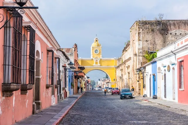 A street in Guatemala with a golden arch at the end and small homes painted pink and blue and white.